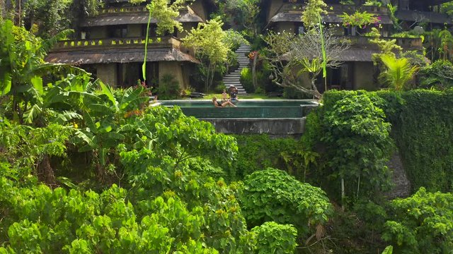 Happy family relaxes in a luxurious infinity pool in a tropical paradise in Ubud, Bali, Indonesia. The drone flies away from the subject high into the sky, revealing a panoramic view. Aerial view 4K.