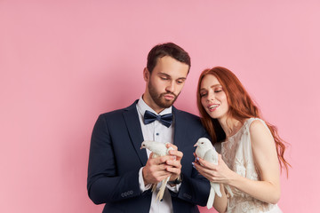 Portrait of caucasian couple wearing marriage wear and holding white pigeons, stand happy isolated over pink background