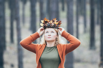 Young Caucasian blonde woman puts on her autumn wreath standing in the pine forest