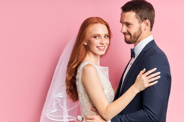 Caucasian man in tuxedo and woman in white wedding dress lovely hug each other and smile, after wedding ceremony posing isolated over pink background.