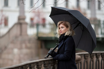 Beautiful young woman in a coat and with an umbrella stands near the fence on the river embankment....