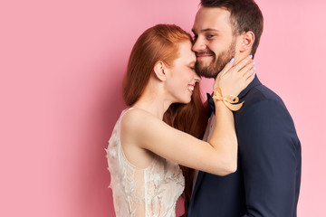 Portrait of redhaired woman in white dress and bearded man in suit hugging each other, isolated pink background