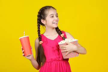 Obraz premium Beautiful young girl holding bucket with popcorn and paper cup on yellow background