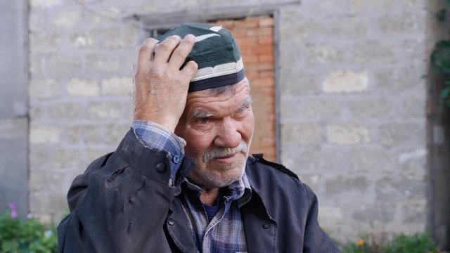 Hoary senior citizen with stubble in shabby navy blue jacket and plaid shirt sitting on street in village. Man stroking his mustache, putting on and removing skullcap, traditional national Muslim hat