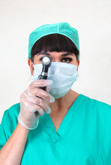 Woman medic holding otoscope to eye. Wearing face mask, hair net and green scrubs. Hand in clear latex glove. Making eye contact. Plain pale background.
