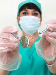 Blurred woman dentist. Focus on dental tools. Photographed from patient point of view. Wearing face mask, green hair net and uniform. Hands in latex gloves. Making eye contact. Plain pale background.