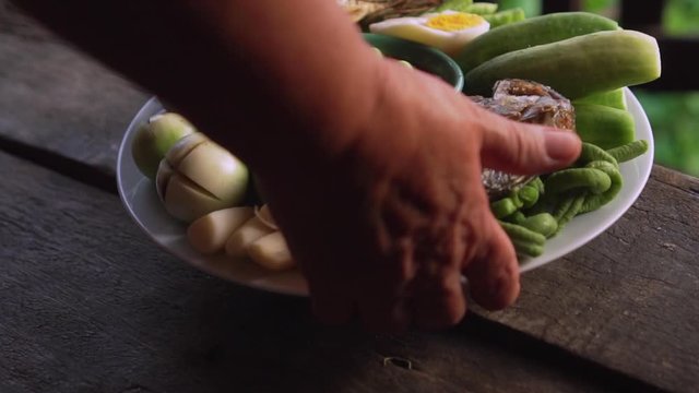 Old asian lady put a plate of fried mackerel with shrimp paste sauce and vegetables on wooden floor. Thai dish. slow motion.