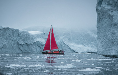 Fototapeta na wymiar A small boat among icebergs. Sailboat cruising among floating icebergs in Disko Bay glacier during midnight sun Ilulissat, Greenland. Studying of a phenomenon of global warming Ices and icebergs