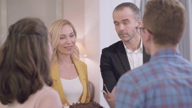 Young Caucasian woman introducing her groom to parents. Severe senior man and his positive blond wife acquainting with their daughter's fiancee. Meeting with parents, acquaintance, family gathering.