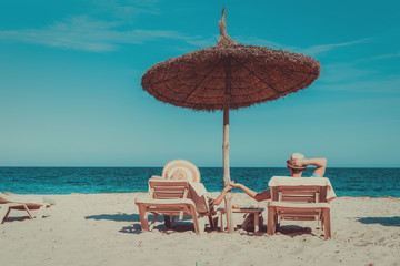 Happy European couple laying on sunbeds under straw umbrella at the tropic beach. They enjoying their vacations and holding hands. Back view