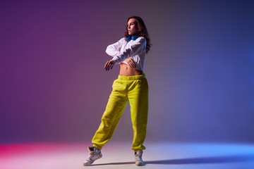 Beautiful young female hip hop dancer with long curly hair, dressed in yellow pants and white roll neck sweater, stands on dance stage studio with naked torso on tiptoe, ready to start dancing