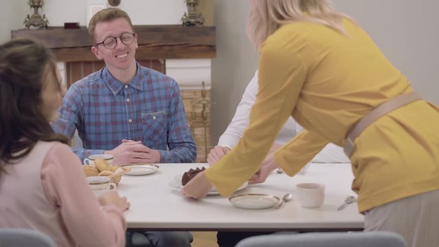 Camera following blond adult Caucasian woman carrying delicious chocolate cake. Mother bringing tasty dessert and putting it at the table. Happy family gathered for dinner.