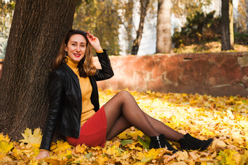 Beautiful young woman in short skirt and leather jacket siting next to the tree in the autumn park....