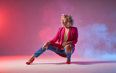Slim tanned woman dressed in stylish clothes, and red heels, sitting on squat, looking away, showing dancing element on studio background, jazz funk street hip hop music