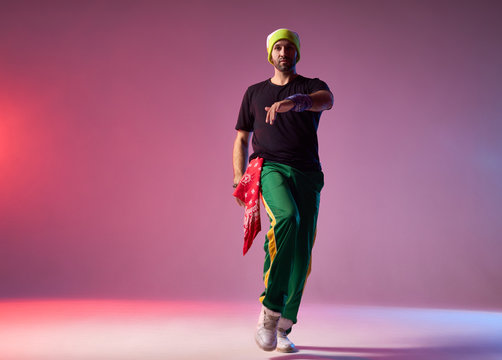 Portrait of dancing man dressed in sportswear and yellow cap, having red bandana demonstrating dancing skills, performing on studio stage, light coloured background