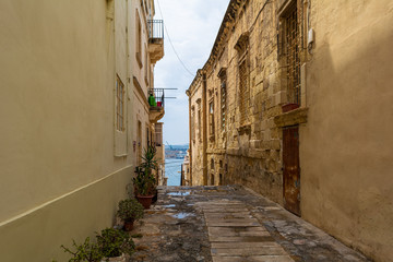 Narrow charming street in Senglea, Malta, with potted plants along the wall and Mediterranean sea seen at the distance.