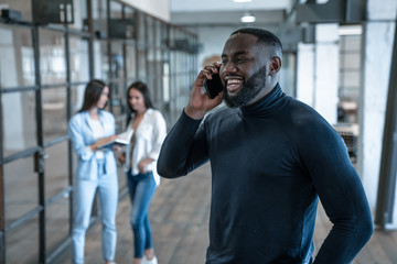 Quick business talk. Handsome young african american smiling man talking on the phone and gesturing while his colleagues standing in the background
