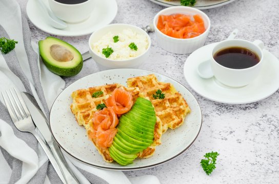 Cheese crispy waffles with cream cheese, smoked salmon and avocado for breakfast with a cup of coffee on a light stone background.