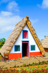 Fototapeta na wymiar Traditional house in Santana, Madeira island, Portugal. Small, wooden, triangular houses with thatched roof and colorful facade. Part of Portuguese heritage. Flower garden in foreground. Sunny day