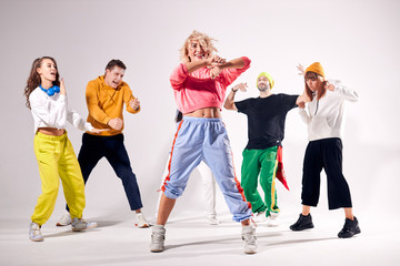 Joyful young blonde girl with long curly hair dressed in sportive clothes, dancing and friends, smiling brightly, raising fists up, showing flat belly with beautiful piercing, in dance studio