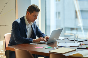 Caucasian businessman wearing a grey smart suit typing on laptop, replying to emails at the office space. Supplies and smartphone scattered around him. Serious and worried expression on his face.