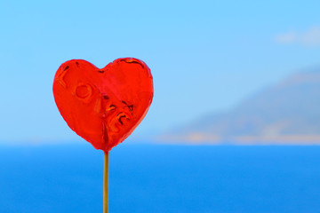  Candy lollipop in the form of a red heart on a background of the blue sea and sky.