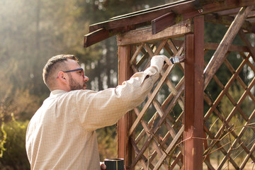 Mężczyzna w ogrodzie maluje na brązowo drewniane deski mebla ogrodowego. Odnawianie i  konserwacja pergoli ogrodowej.