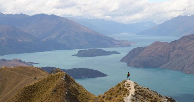 Travel hiker with view of amazing mountain lake nature landscape on New Zealand on famous Wanaka hike to Roys Peak. Happy hiking man jumping having fun in nature on New Zealand. 59.94 FPS SLOW MOTION.