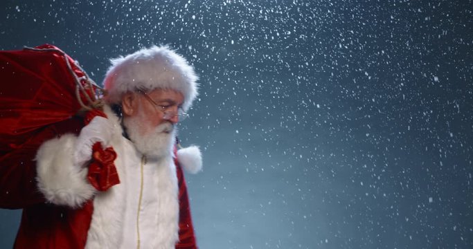 Santa Walking through snow, carrying a bag full of presents, then stopping to look at camera to playfully wink- christmas spirit concept close up 4k footage