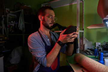 Young cobbler in glasses is working on shoe sole at his dark workplace.