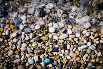 Background of multicolored sea pebbles on the beach in sea foam. Small color wet stones gravel texture background.