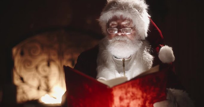Authentic caucasian senior in santa claus clothing looking through book with red cover, sitting near his fireplace - christmas spirit, holidays and celebrations concept close up 4k