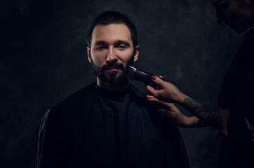 Attractive man have beard trimming with machine at dark barbershop from female.