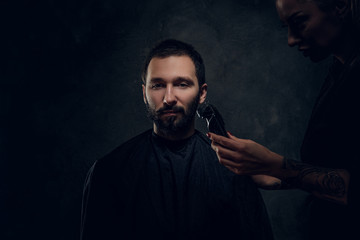 Attractive man have beard trimming with machine at dark barbershop from female.