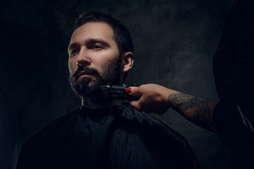 Attractive man have beard trimming with machine at dark barbershop from female.