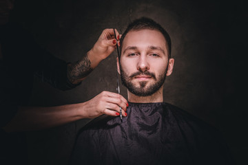 Young gentleman is enjoying mustache and beard trimming at dark barbershop.