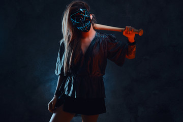 Lawbreaker female in spooky mask is standing at dark studio with bat in her hands.