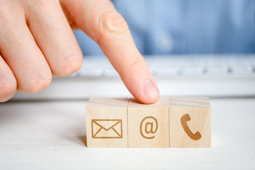 A man points to a wooden cube with the image of an email symbol next to a telephone and a letter with his hand. Contact concept for communication.