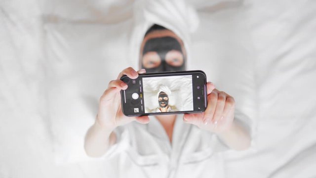 smartphone selfie photo at home wellness, spa, skin care woman with a white towel on her head and black cosmetic mask lying on the bed