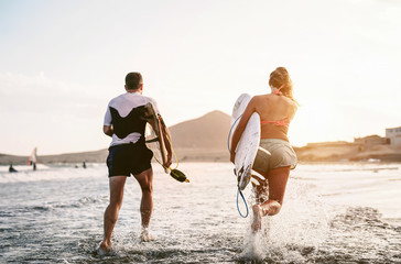 Happy surfers running in the water at sunset time - Young couple having fun surfing in ocean -...