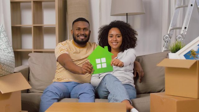 mortgage, eco living and real estate concept - happy african american couple with green paper house and boxes moving to new home and kissing