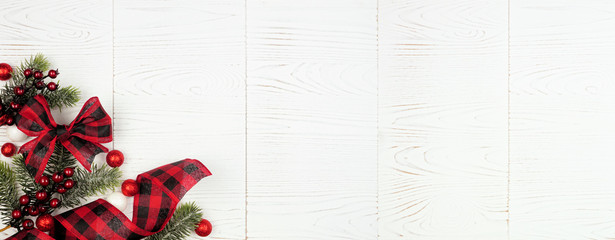 Christmas corner border banner of ornaments, branches and red and black checked buffalo plaid ribbon. Above view on a white wood background.