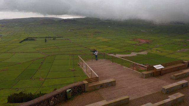 Couple on viewpoint above green farm fields of Terceira island. Woman sits on handrail, msn holds her. Aerial of Azores.