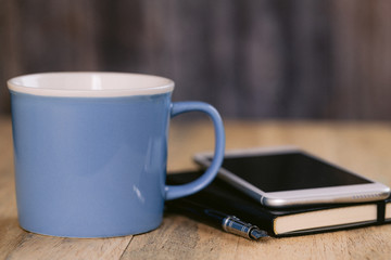 A blue mug with a cellphone and a notebook and pen on a table