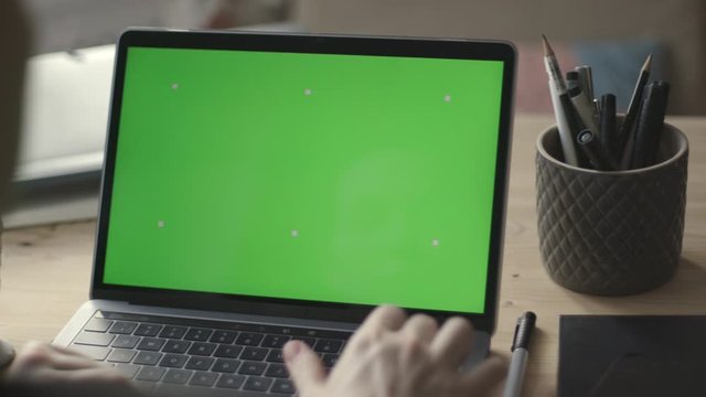 Over the shoulder shot of young woman working on modern laptop with green screen mock-up. Hands on keyboard.