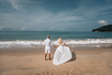 Fototapeta na wymiar Sexy couple in the water.Big sea waves. Beautiful blonde in and a bald man in white clothes are on vacation.Back view. Phuket. Thailand.