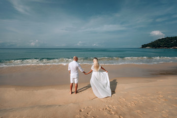Attractive trendy young couple in white clothes are walking along on the beach and holding hands. Beautiful blonde with long hair and a bald man are on vacation. Phuket. Thailand.