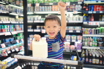 Cute Asian 3 - 4 years old toddler boy child sit in shopping cart choosing milk product in grocery store, Little young kid smiling and raising hand in supermarket, Healthy & Happy child concept