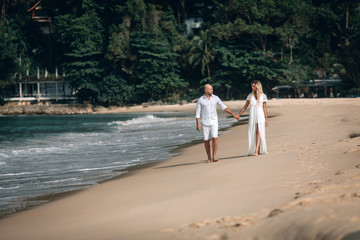 Attractive trendy young couple in white clothes are walking along on the beach and holding hands. Beautiful blonde with long hair and a bald man are on vacation. Phuket. Thailand.