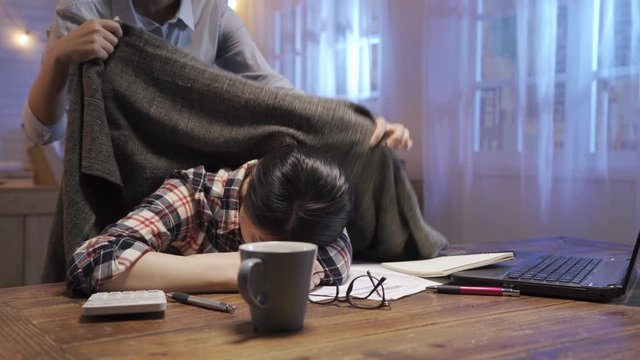 beautiful young asian woman sits at dining table in dark home kitchen at night. businesswoman back from work and tucked roommate in soft blanket in midnight. tired female sleep while doing finance